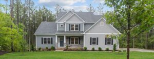 A two-story gray house with white trim and a front porch is surrounded by trees and greenery. There is a well-maintained lawn in front.