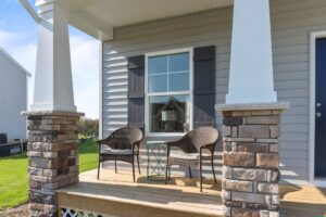 A small front porch with two wicker chairs and a round glass-top table in front of a house with gray siding and white trim. The porch is supported by columns with stone bases.