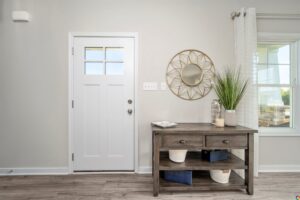 A small wooden console table with two drawers and open shelves stands against a wall next to a front door. It holds decorative items including a plant, lamp, book, and three baskets in an organized manner.