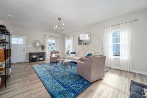 A modern living room with wooden flooring, a blue patterned rug, light-colored furniture, a wall clock, a floor lamp, and large windows with sheer curtains providing ample natural light.