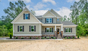 Two-story suburban house with light green and beige siding, dark shutters, a stone entryway, and a front porch. Surrounded by trees and recently landscaped grounds.