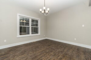 An empty room with light gray walls, a window on the left, and a five-bulb chandelier. The floor is dark wood, and there are white baseboards.
