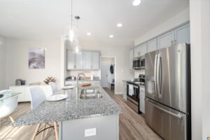 Modern kitchen with granite countertops, stainless steel appliances, light gray cabinets, and an island with a sink. The room features pendant lighting and light wood flooring.