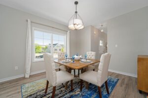 A dining room with a wooden table set for four with blue glasses and napkins. There are four upholstered chairs around the table, and the room has a large window and a modern light fixture.