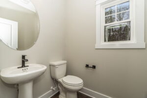 Small bathroom with a pedestal sink, round mirror, and white toilet next to a window. Walls are off-white and the floor is dark wood.