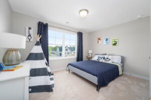 A child’s bedroom with a bed, a striped teepee, a lamp on a white dresser, and three framed pictures on the wall. The room has a large window with navy curtains and carpeted flooring.