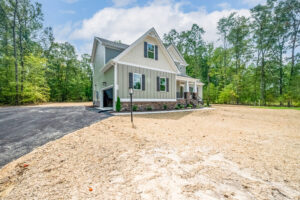 A two-story house with light green siding and a front porch, surrounded by trees and a newly paved driveway.