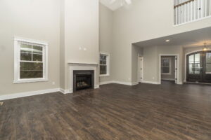 Spacious, empty living room with dark wood flooring, a high ceiling, white walls, and a fireplace. A staircase and doorway are visible in the background.