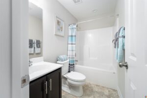 A clean bathroom with a white bathtub and shower curtain, a toilet, a sink with a dark cabinet, and towels hung on the wall. A framed picture hangs on the wall above the towels.