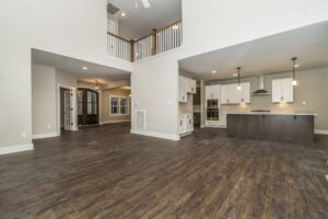 Spacious open-concept living area with wooden floors, featuring a modern kitchen with white cabinetry, an island, and pendant lighting under a high ceiling. Balcony visible on the upper floor.