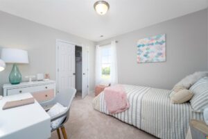 A bedroom with a bed covered in striped linens, a desk with a lamp and chair, art on the walls, and a window with a view of greenery. The room is lit by a ceiling light.