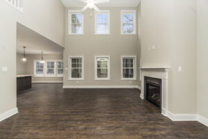 Spacious living area with high ceiling, large windows, wooden floors, and a fireplace. Dining area visible in the background. Walls painted in neutral tones.