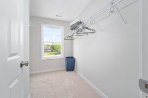 An empty walk-in closet with beige carpet, white walls, wire shelving with several hangers, a blue laundry basket, and a window providing natural light.