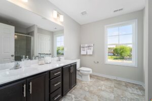 Modern bathroom with a double sink vanity, dark cabinets, a large mirror, towel rack, toilet, and a walk-in shower. The room has beige walls, tiled floors, and a window providing natural light.