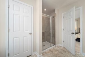 A bathroom featuring a glass-door shower with tile walls, a closed door on the left, and a partially open door on the right. The floor is covered with light-colored tiles.