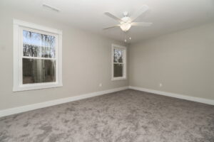 An empty room with beige walls, two windows with a view of trees, a ceiling fan with light, and gray carpet flooring.