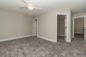 Empty carpeted room with beige walls, a ceiling fan, and two open doorways leading to a closet and hallway.