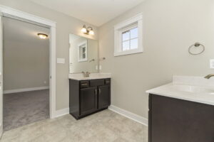 A bathroom with a vanity, sink, mirror, and light fixture. Doorway leads to a carpeted room. Walls and tiles are in neutral colors.