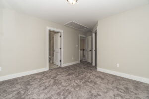 Empty room with beige walls, gray carpet, and three doorways. One door leads to a bathroom; another appears to open to a hallway. Ceiling light fixture is visible.