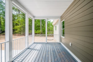 A covered porch with wooden flooring and white railings, overlooking a grassy area bordered by trees. The side of a house with two windows is visible.