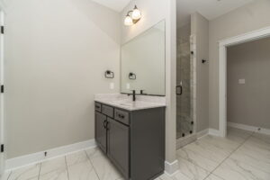 A modern bathroom with a gray vanity, large mirror, dual lights, and a glass-enclosed shower. The floor features white tiles, and the walls are light gray.