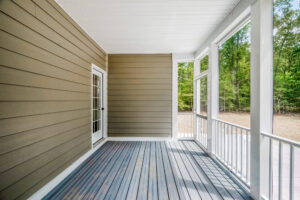 A screened-in porch with white railings, brown siding, and a French door. The floor is made of light gray wood, and the surrounding view shows trees in the background.