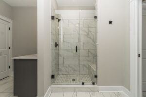 A modern bathroom with a glass-enclosed shower featuring white and gray marble tiles, black fixtures, and a small step. Walls and floor have a similar design.