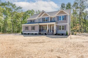 Two-story gray house with a large porch, surrounded by trees, on a spacious plot with a gravel path leading to the entrance.