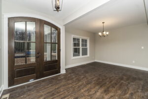 A room with wooden double doors, a window, and a dark wood floor. A chandelier hangs from the ceiling. Walls are light grey.