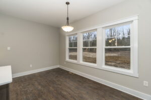 A vacant room with wooden floor, beige walls, a ceiling light, and three large windows overlooking a wooded area.
