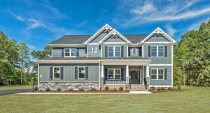 A large two-story gray house with white trim, featuring a front porch and brick steps, surrounded by a grassy yard and trees under a blue sky.