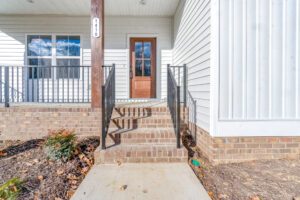 Front porch of a house with white siding and brick steps leading to a wooden door. Black metal railings are on both sides of the steps. A small shrub is on the left.