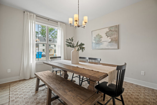Nestled in the heart of Haley Meadows, this dining room features a wooden table, black chairs, and a cozy bench. A captivating painting graces the wall beside a window adorned with white curtains. Above it all, an elegant chandelier casts its warm glow.