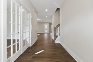 Spacious hallway with wooden flooring, glass-paneled double doors on the left, and a staircase on the right leading to an upper floor. A door is visible at the far end.