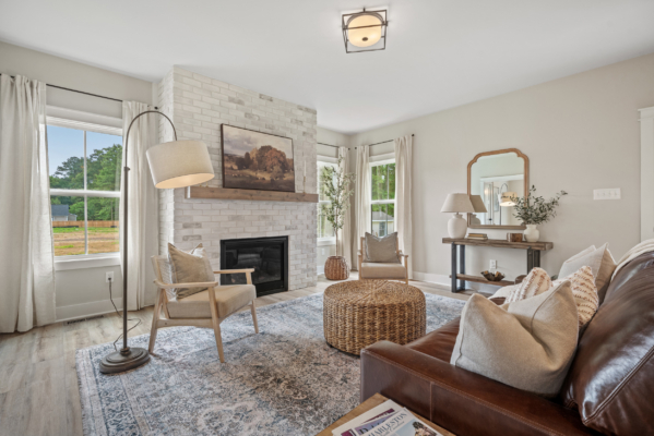 The cozy living room at Haley Meadows features a brick fireplace, a brown sofa, and a round wicker table, all accented by large windows.