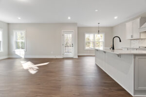Spacious, empty kitchen with light gray walls, wooden floor, white cabinets, a black faucet, pendant light, and glass door leading outside.