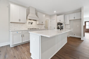 Modern kitchen with white cabinets, subway tile backsplash, and a large marble island. Stainless steel range with hood and wood flooring are visible. Bright natural light fills the space.