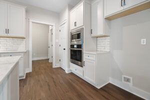 A modern kitchen with white cabinets, stainless steel appliances, and wooden flooring. Features a double oven and tiled backsplash.