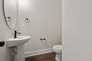 Small bathroom with a pedestal sink, oval mirror, black faucet, toilet, and towel ring. White walls and dark wood flooring.