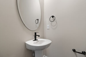 A minimalist bathroom with a round mirror above a white pedestal sink and a black faucet. There is a black towel ring on the wall.