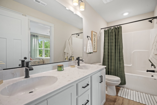 The stylish bathroom in Haley Meadows features double sinks, sleek white countertops, black fixtures, a vibrant green shower curtain, and an abundance of natural light.