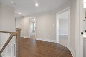 A hallway with wooden floors, beige walls, and white trim, leading to carpeted rooms. Doors on both sides are open, with one brightly lit bathroom visible.