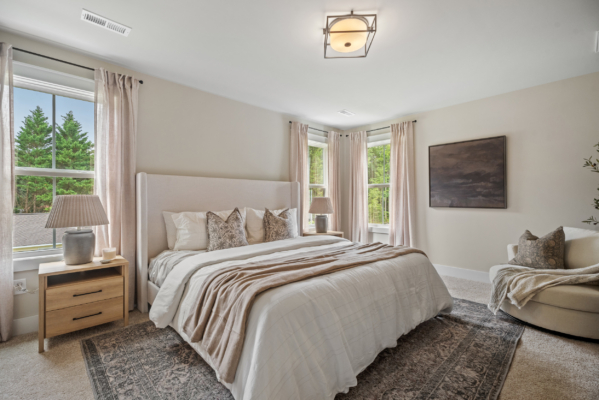 Spacious bedroom in Haley Meadows featuring a large bed, beige bedding, two windows, and a cozy armchair in the corner.