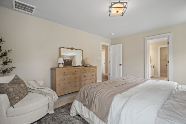 The cozy bedroom in Haley Meadows features a bed, armchair, dresser, and mirror. An open door leads to the bathroom, all wrapped in neutral tones and soft light.