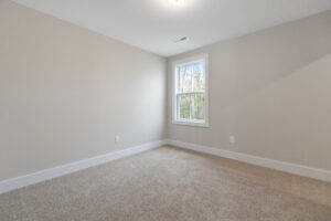 An empty room with beige walls, white trim, and a window. The floor is carpeted, and there's a ceiling light.