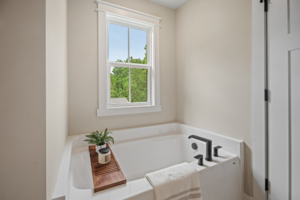 The modern bathroom in Haley Meadows boasts a white tub with sleek black fixtures, accompanied by a cozy towel. A wooden tray holding a vibrant plant sits gracefully by the window, creating an inviting and serene atmosphere.