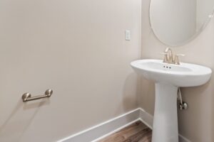 A white pedestal sink with a gold faucet is in a bathroom with a round mirror and wall-mounted towel bar. The floor has wood-style tiles.