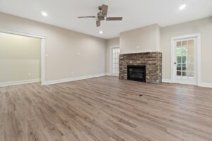 Spacious empty room with hardwood floors, a ceiling fan, stone fireplace, and French doors leading outside. Walls are painted light beige.