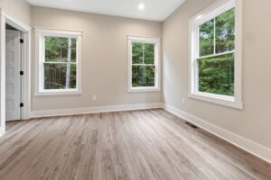 An empty room with hardwood floors, beige walls, and two windows overlooking a wooded area.