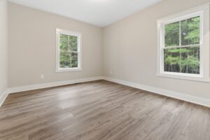 Empty room with light wood flooring, beige walls, and two windows showing a view of green trees.